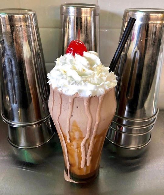 A shake with whipped cream and a cherry, between the metal mixing cups at the fountain.